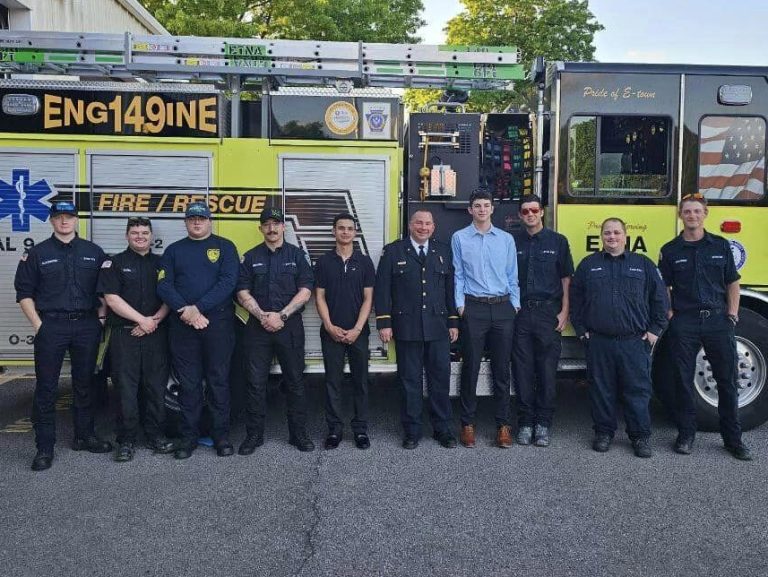 A group of firefighters stands in front of a yellow fire truck labeled "ENG149INE." The group includes both uniformed firefighters, some wearing caps, and others in civilian clothing. They are smiling and pose in front of a large, open garage with trees in the background.