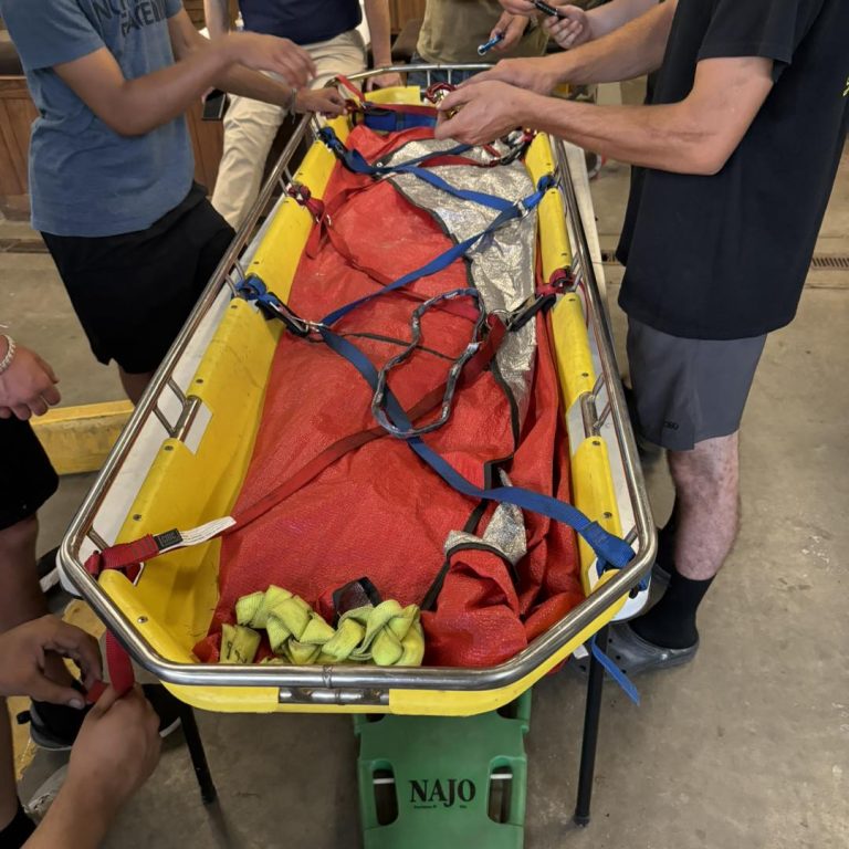 A group of individuals perform a rescue training exercise, securing a stretcher with red and silver straps. The stretcher is yellow and elevated on a green stand labeled "NAJO." The participants' hands are visible, working to adjust and tighten the straps.