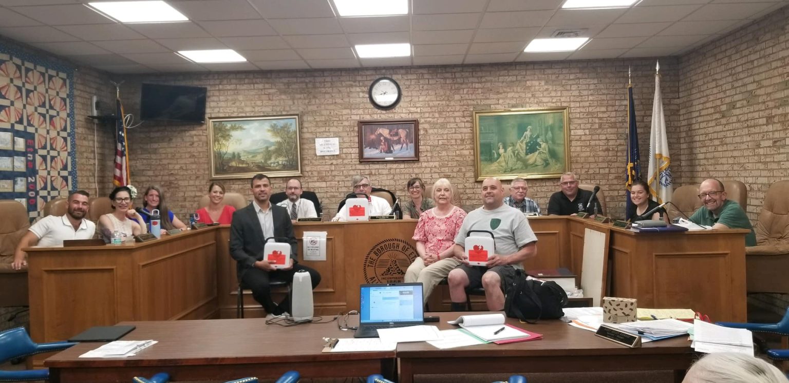 A group of people sitting and standing in a room that resembles a council chamber. There are paintings on the brick walls behind them, flags in the corners, and several tables with papers and laptops. Some individuals hold items with a red cross logo.