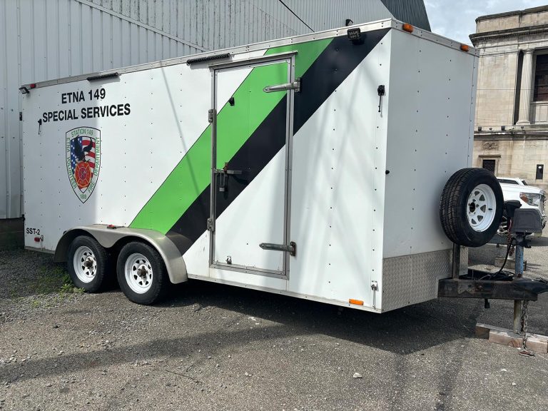 A white utility trailer with "ETNA 149 SPECIAL SERVICES" and a colorful emblem on the side, along with a green and black diagonal stripe. It is parked next to a building with the spare tire mounted on the front. The ground is gravelly with some grass.