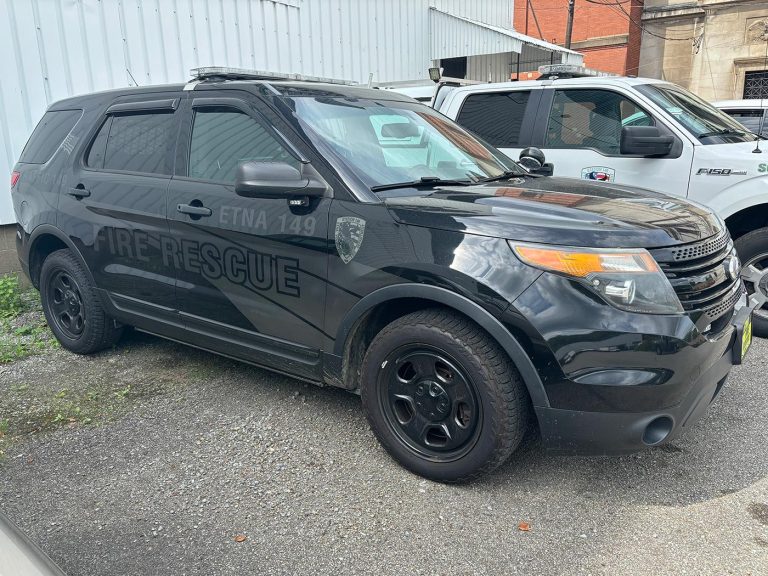 A black SUV marked with "Fire Rescue" and "ETNA 749" on the side is parked on a gravel lot near white buildings. The vehicle has emergency lights on the roof and a shield emblem on the door. A white truck is parked adjacent to the SUV.