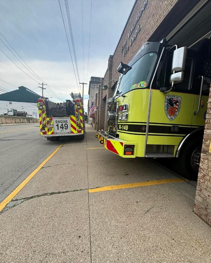 A bright yellow fire truck with the identifier "R-149" is partially parked in a firehouse garage next to another fire truck labeled "ENGINE 149." Both trucks have emergency lights and reflective markings. Brick buildings and utility poles line the adjacent street.