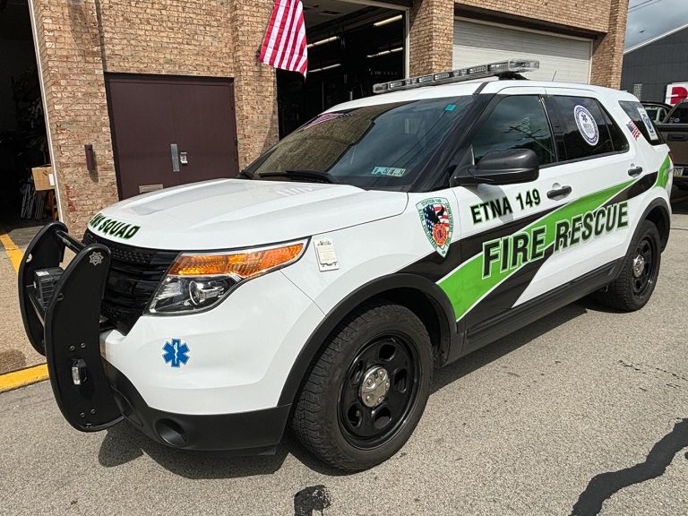 A white and green fire rescue SUV is parked in front of a brick building with an American flag visible to the left. The vehicle has a push bumper, emergency lights, and "Etna 149" markings. The SUV is branded with "Fire Rescue" and has medical and fire department insignias.